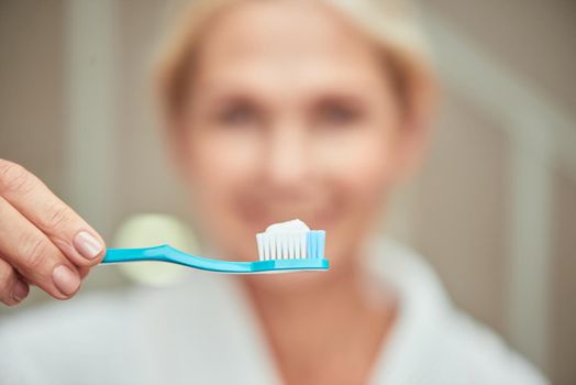 Toothbrush with white paste in hand on blurred woman in spa background, selective focus. Beauty, skincare and cosmetology concept
