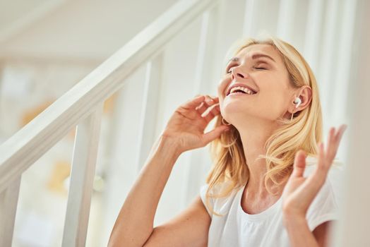 Happy middle aged caucasian woman enjoying music using wireless earphone while sitting indoors with raised hands and closed eyes. People spending time at home