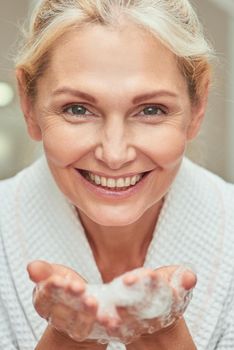 Close up portrait of happy middle aged caucasian woman with open palms full of foam looking at camera and smiling at spa. Beauty, skincare and cosmetology concept
