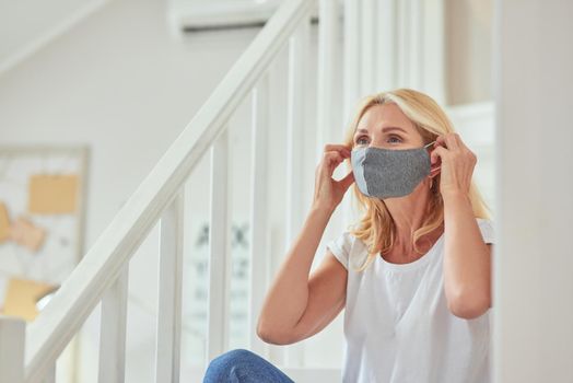 Middle aged caucasian woman putting protective mask on face sitting at modern apartment alone. Isolation at home concept