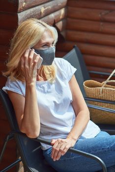 Middle aged caucasian woman in protective mask sitting in chair on balcony looking sideways while talking on phone, vertical shot. Isolation at home concept