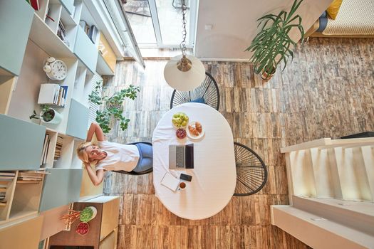 Top view on blonde middle aged caucasian business woman resting on chair with head up while working at home sitting near kitchen table with laptop and smartphone. Working from home concept