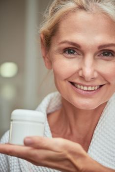 Beautiful middle aged caucasian woman with clean shiny skin holding skin care cream bottle on palm while smiling at camera, selective focus. Beauty, skincare and cosmetology concept