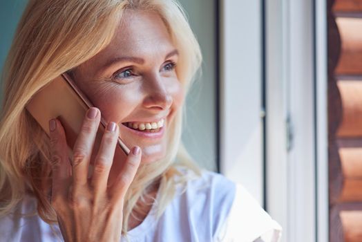 Portrait of middle aged caucasian woman looking outside to balcony standing indoors with phone near ear. People spending time at home