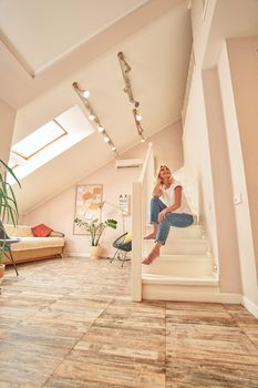 Happy middle aged caucasian woman sitting with raised head on stairs in light flat, bottom view. People spending time at home