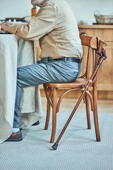 Cropped photo of senior man sitting at the table on the kitchen with cane near him. Care and health concept