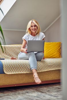 Middle aged caucasian woman smiling at camera while working on laptop sitting on comfortable sofa. Working from home, freelance concept