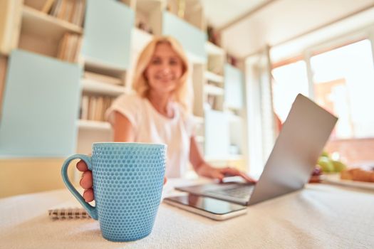 Light blue cup on kitchen table in front of working middle aged caucasian woman, selective focus. Working from home concept