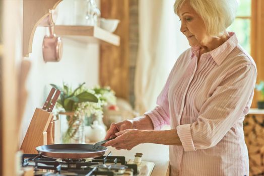 Side view of smiling senior woman going to bake pancakes for breakfast at home. Domestic lifestyle concept