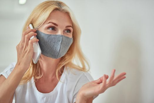 Middle aged caucasian woman with protective mask looking sideways and gesturing while talking on phone sitting at home. Isolation at home concept