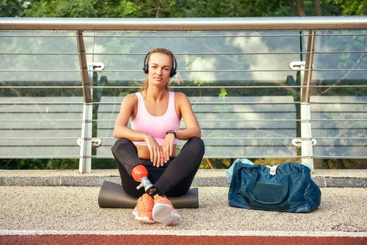 Confident in myself. Portrait of young woman in sports clothing and headphones with leg prosthesis sitting on the bridge and looking at camera. Disabled sport concept. Motivation. Healthy lifestyle