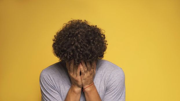 Young man covers his face with his palms. Curly guy in a gray t-shirt, isolated on a yellow background. Stress concept.