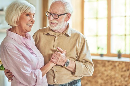 Happy senior man in glasses holding his wife hand while looking at her at home with window on the background. Lifestyle concept