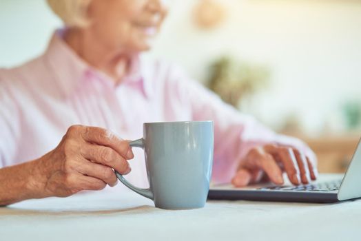 Cropped photo of happy senior woman typing on laptop while enjoying hot drink at home. Domestic lifestyle concept