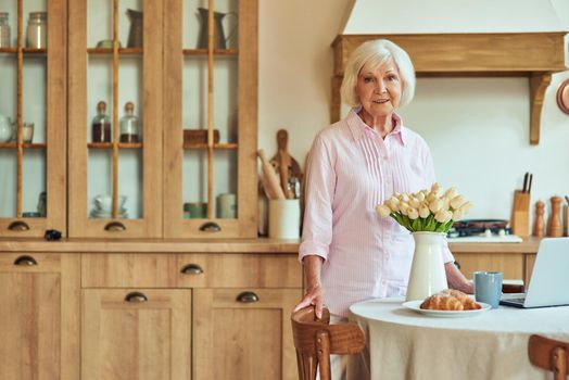 Smiling elderly lady near table with laptop, cup of tea and croissant at home during morning breakfast. Domestic lifestyle concept