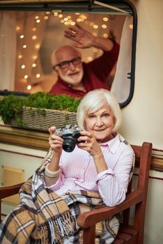 Happy elderly woman resting on the porch of motorhome and holding camera while her husband waving through the window. Travel concept