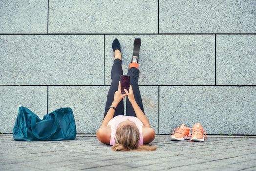 Relaxing after training. Top view of woman with leg prosthesis in sports clothing holding her smartphone while sitting outdoors. Disabled sport concept. Motivation. Digital