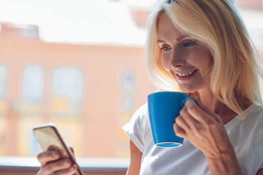 Pretty middle aged caucasian woman with tea cup and smartphone indoors near big window. People spending time at home