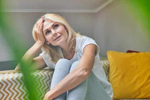 Middle aged caucasian woman dreaming while sitting with raised head on comfortable sofa in her flat, viewed through green plant. People spending time at home