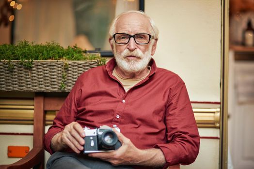 Waist up of senior gray-haired man sitting on wooden chair and holding camera on the porch of the motorhome. Travel concept