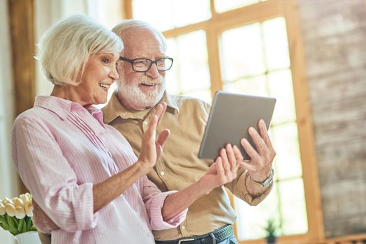 Waist up of smiling senior man and woman waving at screen while holding digital tablet and chatting during video call. Lifestyle concept
