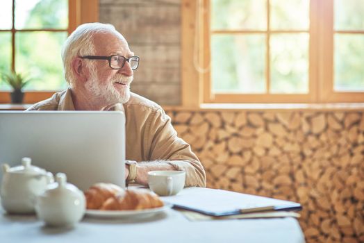 Happy elderly man sitting at the table in living room while using laptop in his home with window on the background. Lifestyle concept