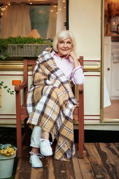 Full length photo of beautiful senior woman with a plaid sitting on a wooden chair near her motorhome. Travel concept
