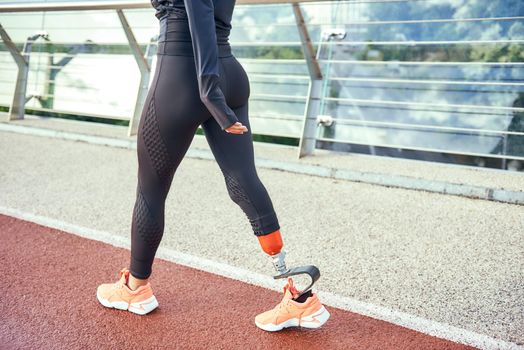 Cropped photo of disabled athlete woman with prosthetic leg in sportswear walking on the bridge. Disabled sport concept. Motivation. Healthy lifestyle