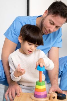 Young therapist helping cute little boy who has cerebral palsy, playing with developing toy at rehabilitation clinic. High quality photo