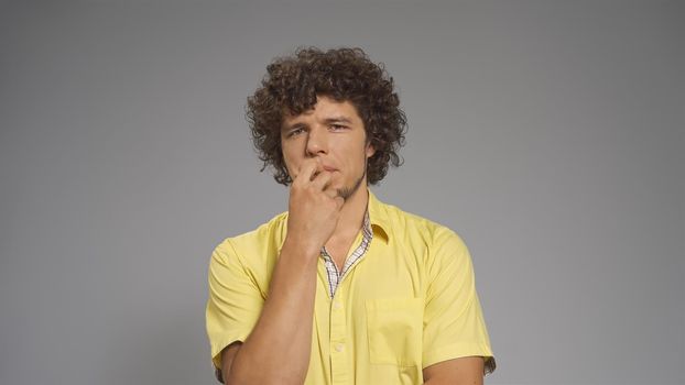 Pensive young man doubts covering his mouth with hand. Cut out on gray background.