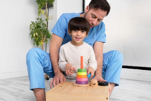 Young therapist helping cute little boy who has cerebral palsy, playing with developing toy at rehabilitation clinic. High quality photo