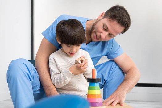 Young therapist helping cute little boy who has cerebral palsy, playing with developing toy at rehabilitation clinic. High quality photo