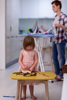 happy family spending time together at home  cute little daughter in a pink dress playing and painting the jewelry box while young redhead mother ironing clothes behind her