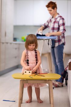 happy family spending time together at home  cute little daughter in a pink dress playing and painting the jewelry box while young redhead mother ironing clothes behind her