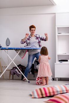 happy family having fun together at home  cute little daughter in a pink dress playing and dancing while young redhead mother ironing clothes behind her