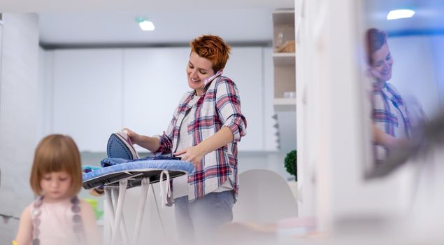 happy family spending time together at home  cute little daughter in a pink dress playing and painting the jewelry box while young redhead mother ironing clothes behind her