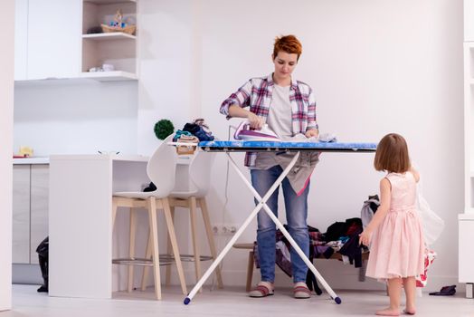 happy family having fun together at home  cute little daughter in a pink dress playing and dancing while young redhead mother ironing clothes behind her