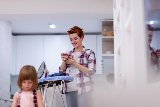 happy family spending time together at home  cute little daughter in a pink dress playing and painting the jewelry box while young redhead mother ironing clothes behind her