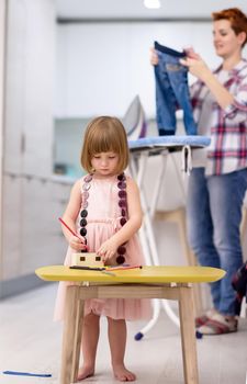 happy family spending time together at home  cute little daughter in a pink dress playing and painting the jewelry box while young redhead mother ironing clothes behind her