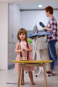 happy family spending time together at home  cute little daughter in a pink dress playing and painting the jewelry box while young redhead mother ironing clothes behind her