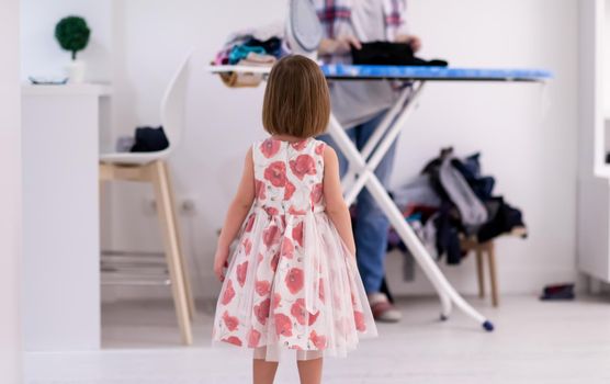 happy family having fun together at home  cute little daughter in a pink dress playing and dancing while young redhead mother ironing clothes behind her