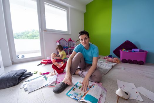 happy family spending time together.young mother and her cute llittle daughter enjoying free time on the floor at home.mom reading a magazine while her daughter playing with dolls in the background near the window