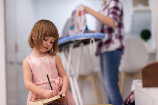 happy family spending time together at home  cute little daughter in a pink dress playing and painting the jewelry box while young redhead mother ironing clothes behind her