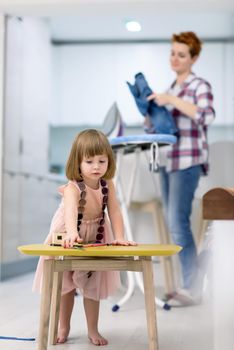 happy family spending time together at home  cute little daughter in a pink dress playing and painting the jewelry box while young redhead mother ironing clothes behind her