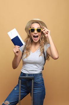 Cheerful blonde woman posing with baggage and holding passport with tickets while looking away over beige background
