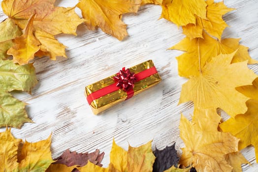 Flat lay cute composition with gift box and yellow autumn leaves. Holiday present decorated red ribbon bow on vintage wooden desk. Happy thanksgiving congratulation. Still life of autumn season.