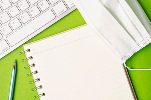 Open blank diary and keyboard computer with disposable face mask on green background.