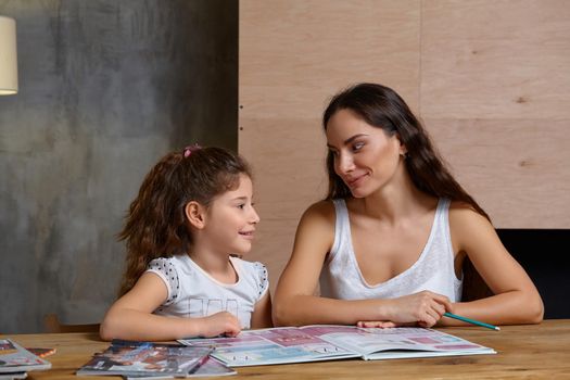 Portrait of a mother helping her small sweet and cute daughter to make her homework indoors, looking at each other and smiling.