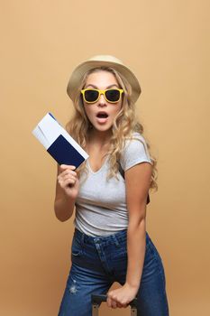 Cheerful blonde woman in sunglasses posing with baggage and holding passport with tickets over beige background