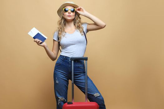 Cheerful blonde woman in sunglasses posing with baggage and holding passport with tickets over beige background